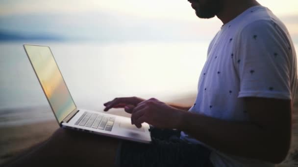 Молодой, красивый мужчина, работающий над ноутбуком на пляже на закате солнца. Молодой бизнесмен работает на тропическом острове на закате. Фрилансер работает на берегу моря.
. - Кадры, видео
