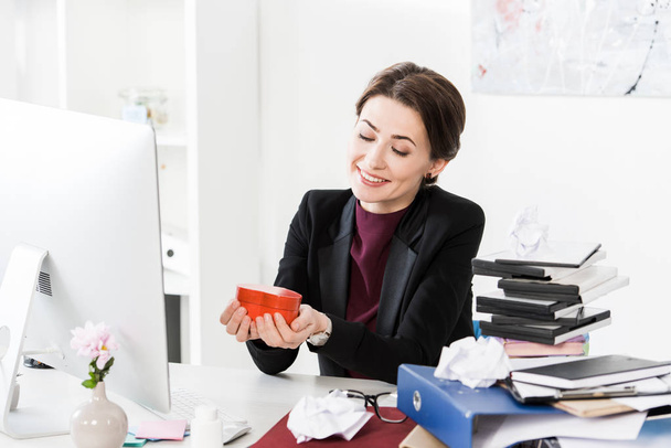 ευτυχισμένη ελκυστικός επιχειρηματίας κρατώντας καρδιά σχήμα κόκκινο κουτί παρόντες στο γραφείο - Φωτογραφία, εικόνα