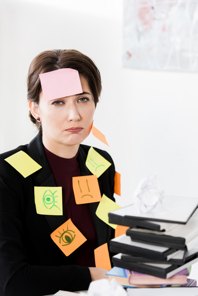 расстроен привлекательный бизнес-женщина сидит с наклейками на лице и одежде в офисе, глядя на камеру
 - Фото, изображение