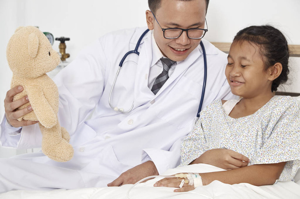 Медицинская концепция. Доктор, держащий плюшевого мишку с маленькой девочкой, счастливо улыбается в больнице, осматриваясь.
 - Фото, изображение