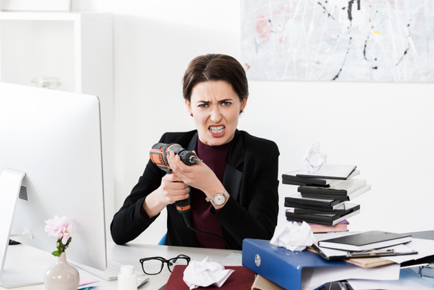 агрессивная привлекательная деловая женщина, проводящая электрическую дрель в офисе и смотрящая в камеру
 - Фото, изображение