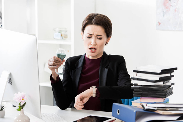 привлекательная шокированная деловая женщина держит песочные часы и проверяет время с наручными часами в офисе
 - Фото, изображение