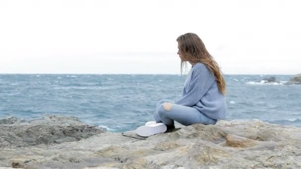Visão lateral retrato de um adolescente triste sentado na praia em um dia ventoso
 - Filmagem, Vídeo