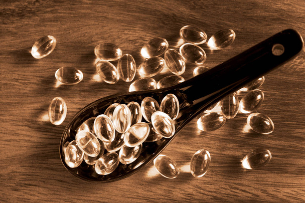 Transparent yellow pills in ceramic spoon on wood background, capsules cod liver oil, omega 3 in black tablespoon. - 写真・画像