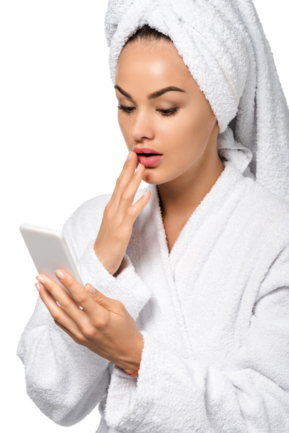 überrascht Mädchen im Bademantel schaut auf Smartphone-Bildschirm isoliert auf weiß - Foto, Bild