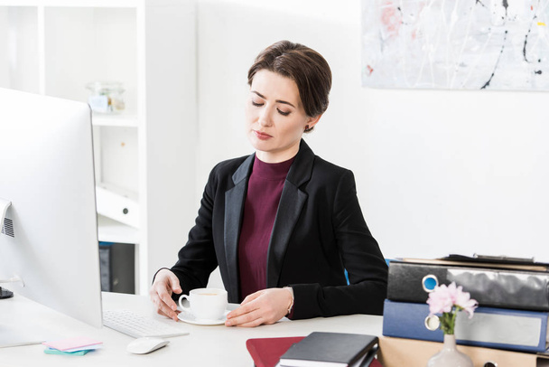 attractive businesswoman holding cup of tea at table in office - Photo, Image