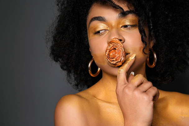 Αφρικανικός-αμερικανική γυναίκα με χρυσή μπογιά στο σώμα της και το λουλούδι στο στόμα με μαύρο φόντο - Φωτογραφία, εικόνα