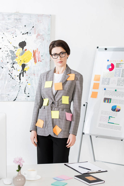aantrekkelijke zakenvrouw met papieren stickers op jas permanent in de buurt van flip-over en kijken naar camera in kantoor - Foto, afbeelding