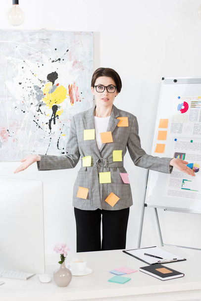 shocked businesswoman with paper stickers on jacket standing near flipchart and showing shrug gesture in office - Фото, изображение