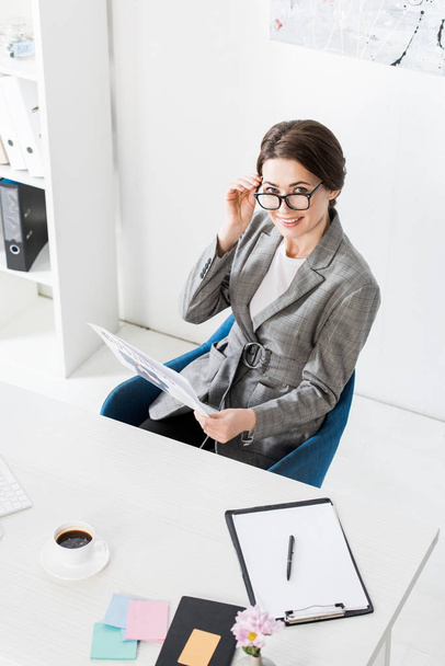 high angle view of smiling attractive businesswoman in glasses holding newspaper in office - Foto, Bild