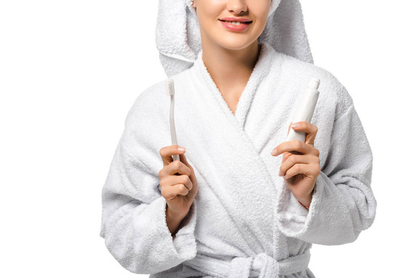 cropped view of girl in bathrobe holding toothbrush and toothpaste and smiling isolated on white - Foto, imagen