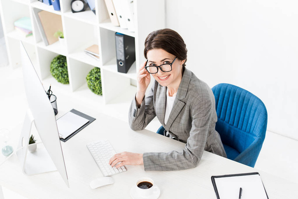 vista de ángulo alto de sonriente atractiva mujer de negocios utilizando el ordenador en la mesa en la oficina y mirando a la cámara
 - Foto, Imagen
