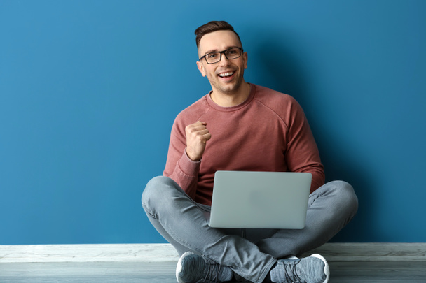 Succesvolle jonge man met laptop zitting in de buurt van kleur muur - Foto, afbeelding