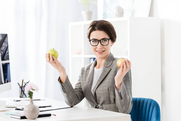attractive businesswoman holding apples and looking at camera in office  - 写真・画像