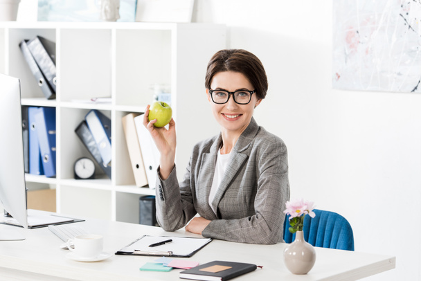 lächelnde attraktive Geschäftsfrau mit Apfel im Büro und Blick in die Kamera - Foto, Bild