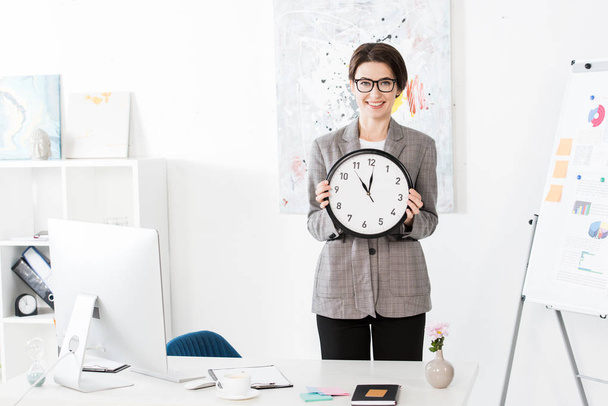 sorridente atraente empresária segurando relógio de parede e olhando para a câmera no escritório
 - Foto, Imagem