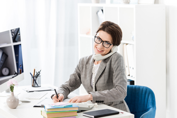 smiling attractive businesswoman talking by stationary telephone in office and looking at camera - Photo, Image