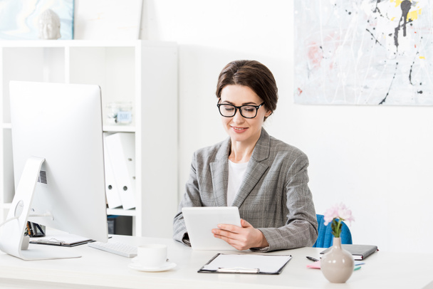 neşeli çekici iş kadını ofiste masada tablet kullanarak gri takım elbise - Fotoğraf, Görsel