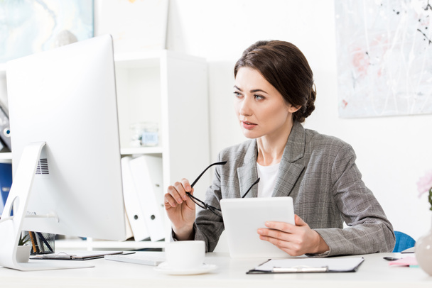 mooie zakenvrouw in grijs pak holding tablet en computer in kantoor kijken - Foto, afbeelding