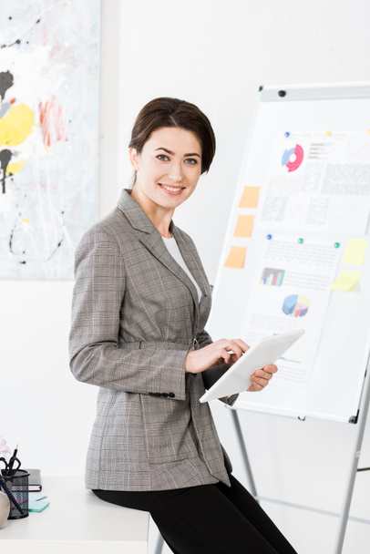 lächelnde attraktive Geschäftsfrau im grauen Anzug sitzt am Tisch und benutzt Tablet im Büro - Foto, Bild