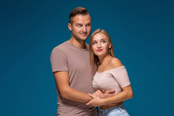 Young couple is hugging on blue background in studio. They wear T-shirts, jeans and smile. - Foto, imagen