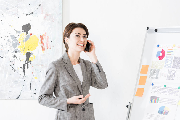 attractive businesswoman in grey suit standing near flipchart and talking by smartphone in office - 写真・画像