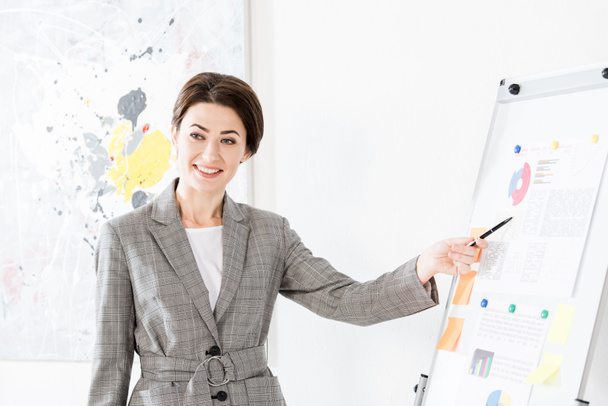 усміхнена красива бізнес-леді в сірому костюмі вказує на фліпчарт під час презентації проекту в офісі
 - Фото, зображення