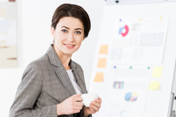 cheerful beautiful businesswoman in grey suit holding cup of coffee in office - Zdjęcie, obraz