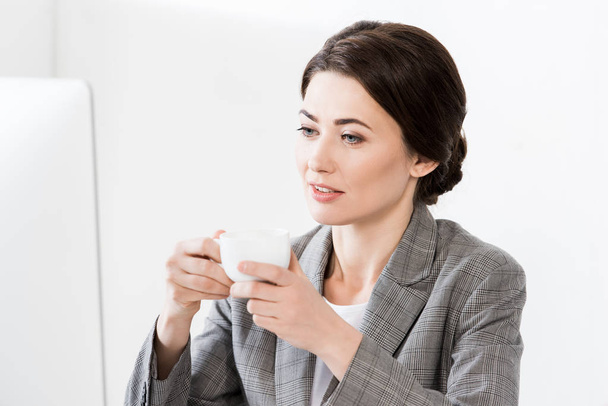 attractive businesswoman in grey suit sitting at table with computer and holding cup of coffee in office - Foto, Imagen