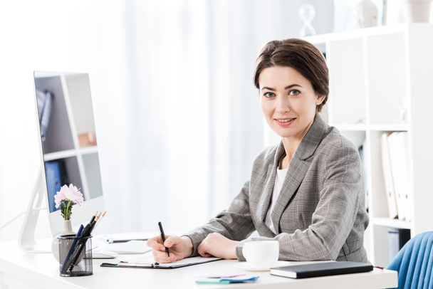 aantrekkelijke zakenvrouw in grijs pak zitten aan tafel met computer, schrijven iets naar het Klembord en kijken naar camera in kantoor - Foto, afbeelding