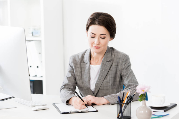 attractive businesswoman in grey suit writing something to clipboard in office - Foto, Imagen