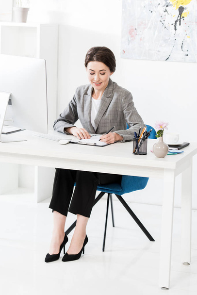 attractive businesswoman in grey suit writing something to clipboard at table in office - Фото, изображение