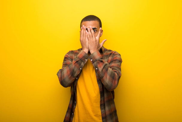 Joven hombre afroamericano sobre vibrante fondo amarillo cubriendo los ojos con las manos y mirando a través de los dedos
 - Foto, imagen