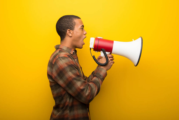 Young african american man on vibrant yellow background shouting through a megaphone to announce something in lateral position - Fotoğraf, Görsel