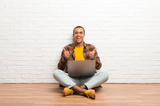 Uomo afroamericano seduto sul pavimento con il suo computer portatile dando un pollice in su gesto con entrambe le mani e sorridente
 - Foto, immagini
