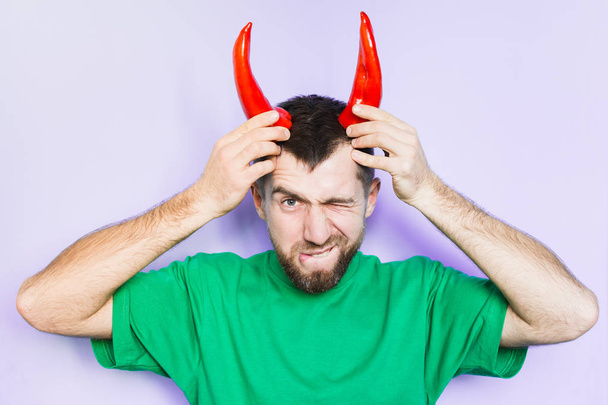 Young man holding red capi peppers above his head which looks like horns, angry and evil face. Light purple background. - Foto, immagini