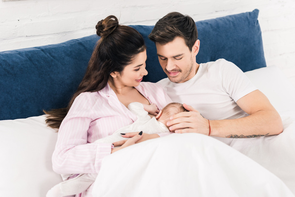 portrait of smiling mother breastfeeding little baby with husband near by on bed at home - Foto, Bild