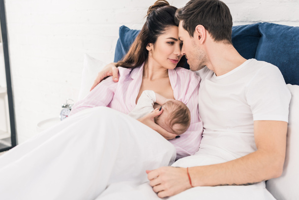 portrait of young mother breastfeeding little baby with husband near by on bed at home - Photo, Image