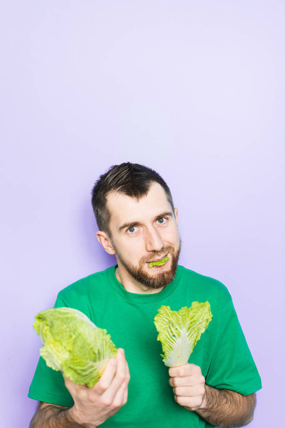 Young man biting on a leaf of Beijing napa cabbage, offering the product to the viewer. Light purple background, copy space, vertical orientation. - Фото, зображення