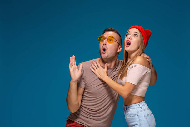 Surprised couple in casual clothes pointing with their index fingers up looking there with great astonishment isolated over blue background - Photo, Image
