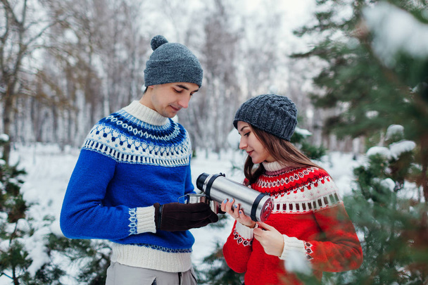 Young couple pouring hot tea out of thermos into cups in winter forest. Happy people relaxing outdoors during holidays - Photo, Image