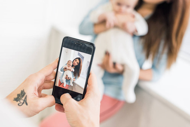 recortado disparo de hombre tomando la foto de la esposa con el bebé en las manos en el teléfono inteligente
 - Foto, imagen
