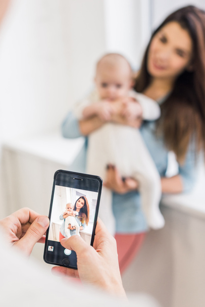 cropped shot of man taking picture of wife with baby in hands on smartphone - 写真・画像