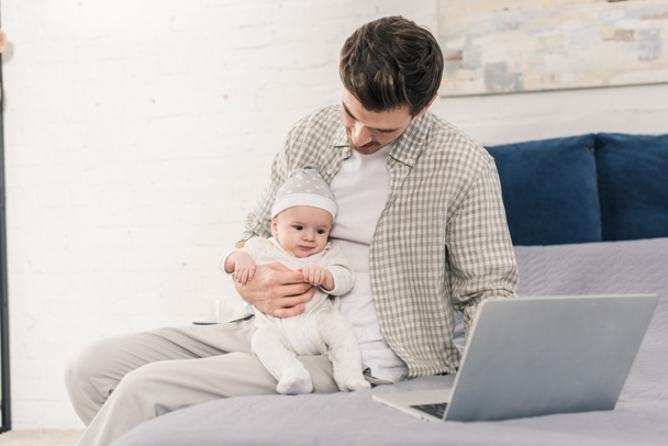 portrait of man working on laptop with little baby in hands at home, work and life balance concept - Fotografie, Obrázek