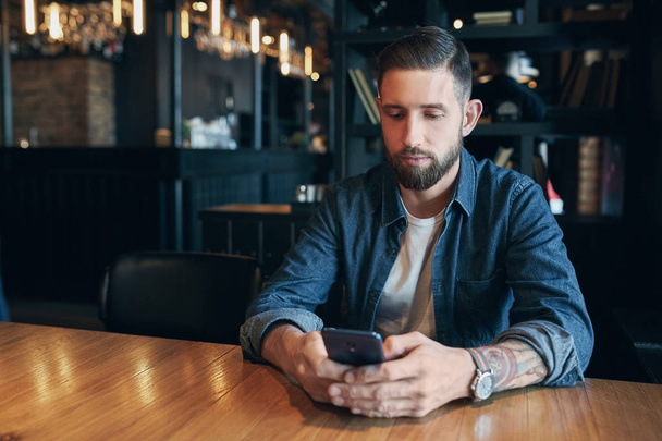 Młody Brodaty biznesmen, ubrany w koszulę denim, siedząc przy stoliku w kawiarni i wykorzystanie smartfonu. Człowiek za pomocą gadżetu. - Zdjęcie, obraz