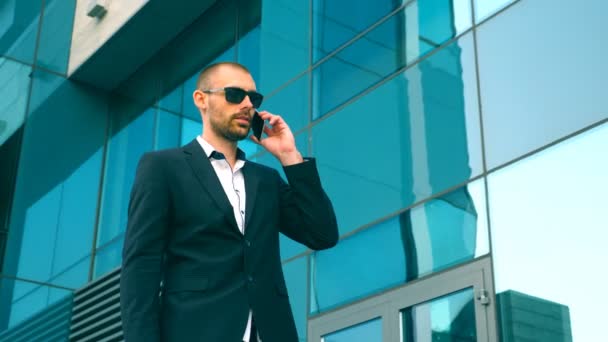 Businessman in sunglasses walking near office and talking on phone. Young happy business man going outdoor and speaking on cellphone. Portrait of guy having work conversation. Slow motion Close up - Footage, Video