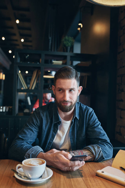 Young bearded businessman,dressed in a denim shirt, sitting at table in cafe and use smartphone. Man using gadget. - Photo, image