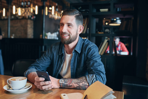 Młody Brodaty biznesmen, ubrany w koszulę denim, siedząc przy stoliku w kawiarni i wykorzystanie smartfonu. Człowiek za pomocą gadżetu. - Zdjęcie, obraz