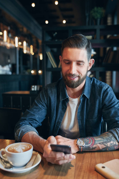 Young bearded businessman,dressed in a denim shirt, sitting at table in cafe and use smartphone. Man using gadget. - Photo, Image