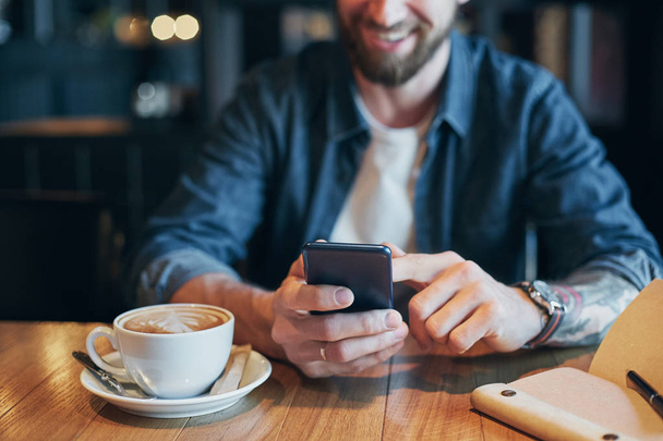 Мужчина руки в джинсовой рубашке слайд с пальцем на экране своего смартфона, возле чашки с кофе на деревянном столе
 - Фото, изображение